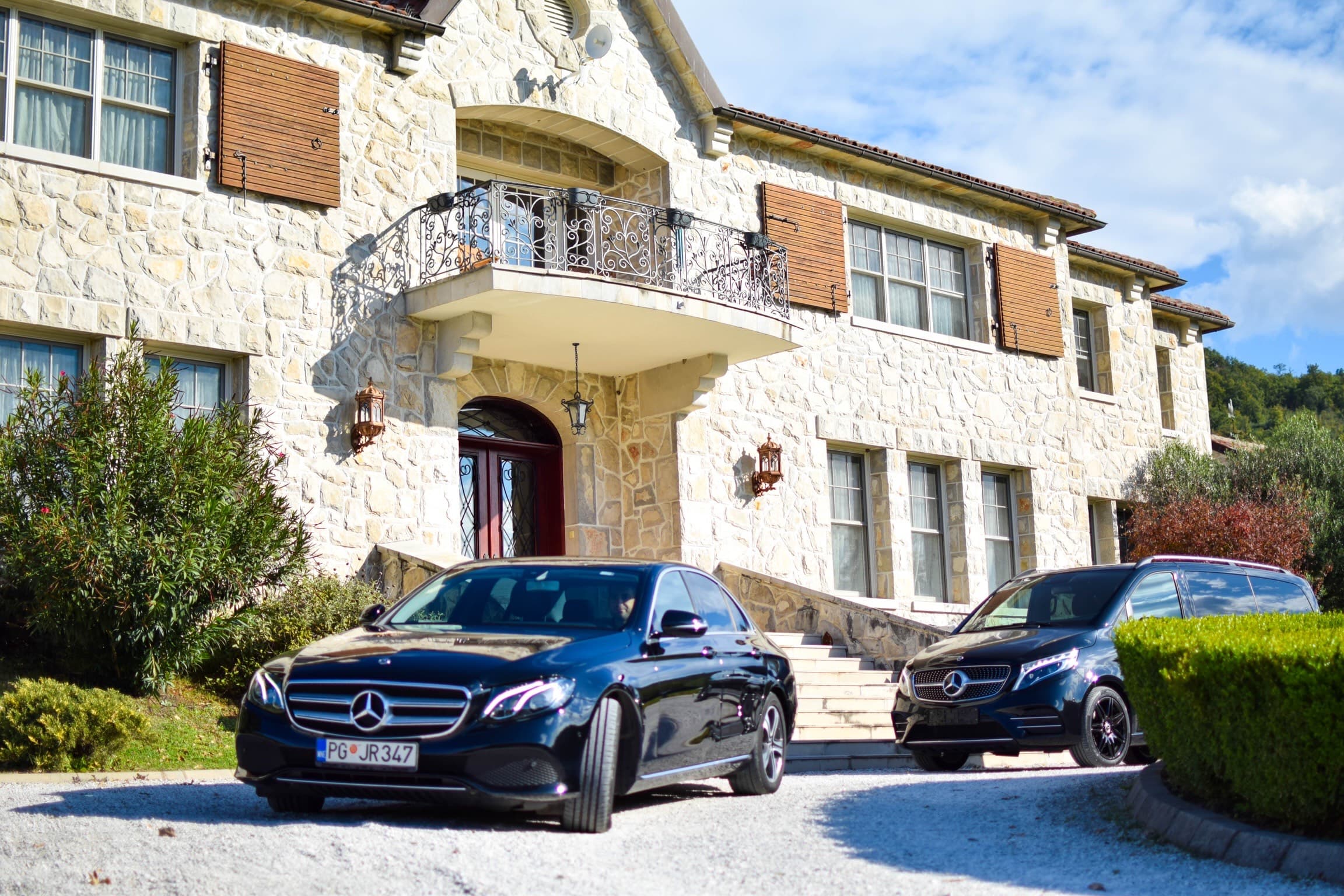 Crni Mercedes-Benz sedan i kombi parkirani ispred kamene kuće sa balkonom