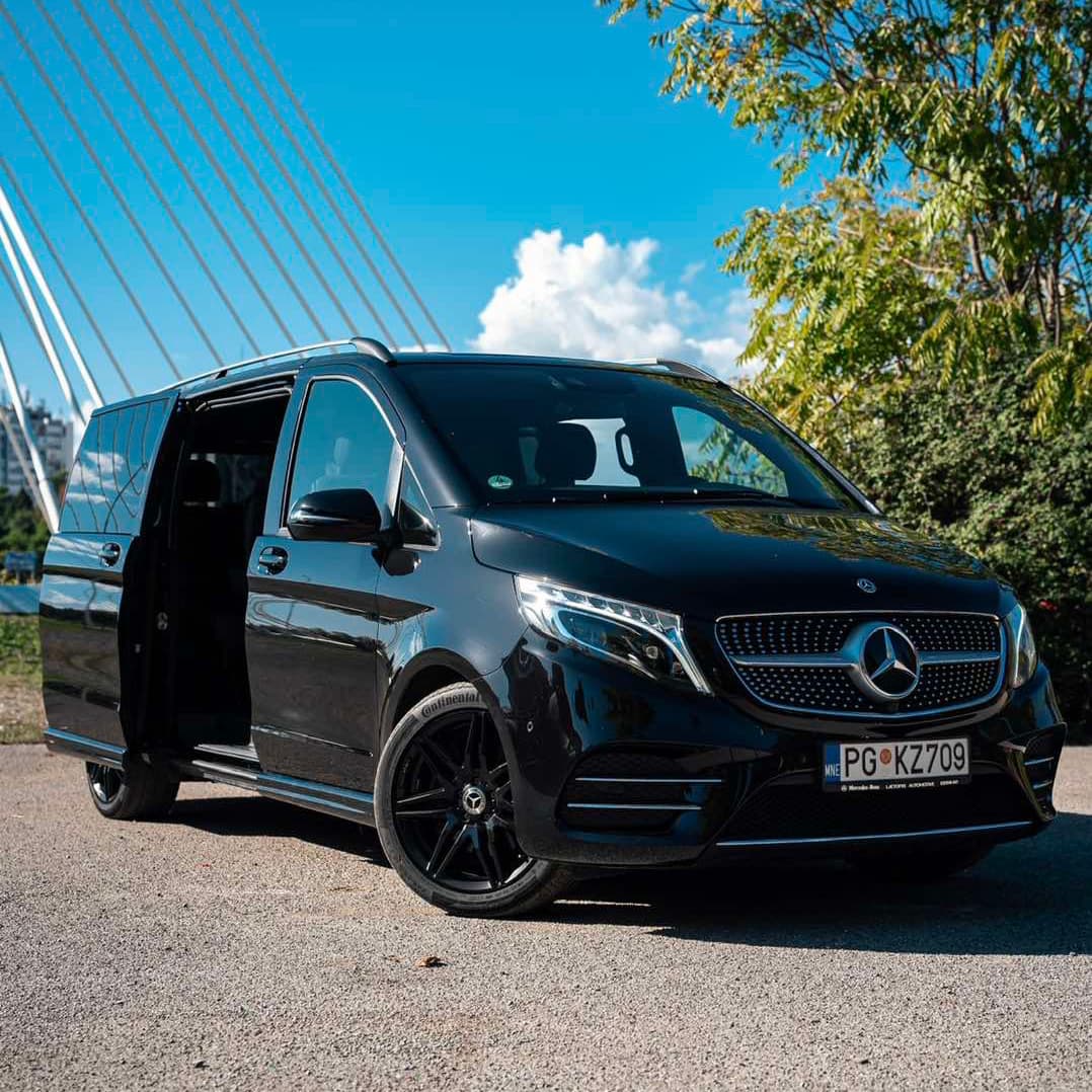 Crni Mercedes-Benz V-Class kombi parkiran sa otvorenim bočnim vratima, prikazujući svoj elegantan dizajn i luksuzni izgled, u blizini mosta Milenijum i čistim plavim nebom u pozadini.